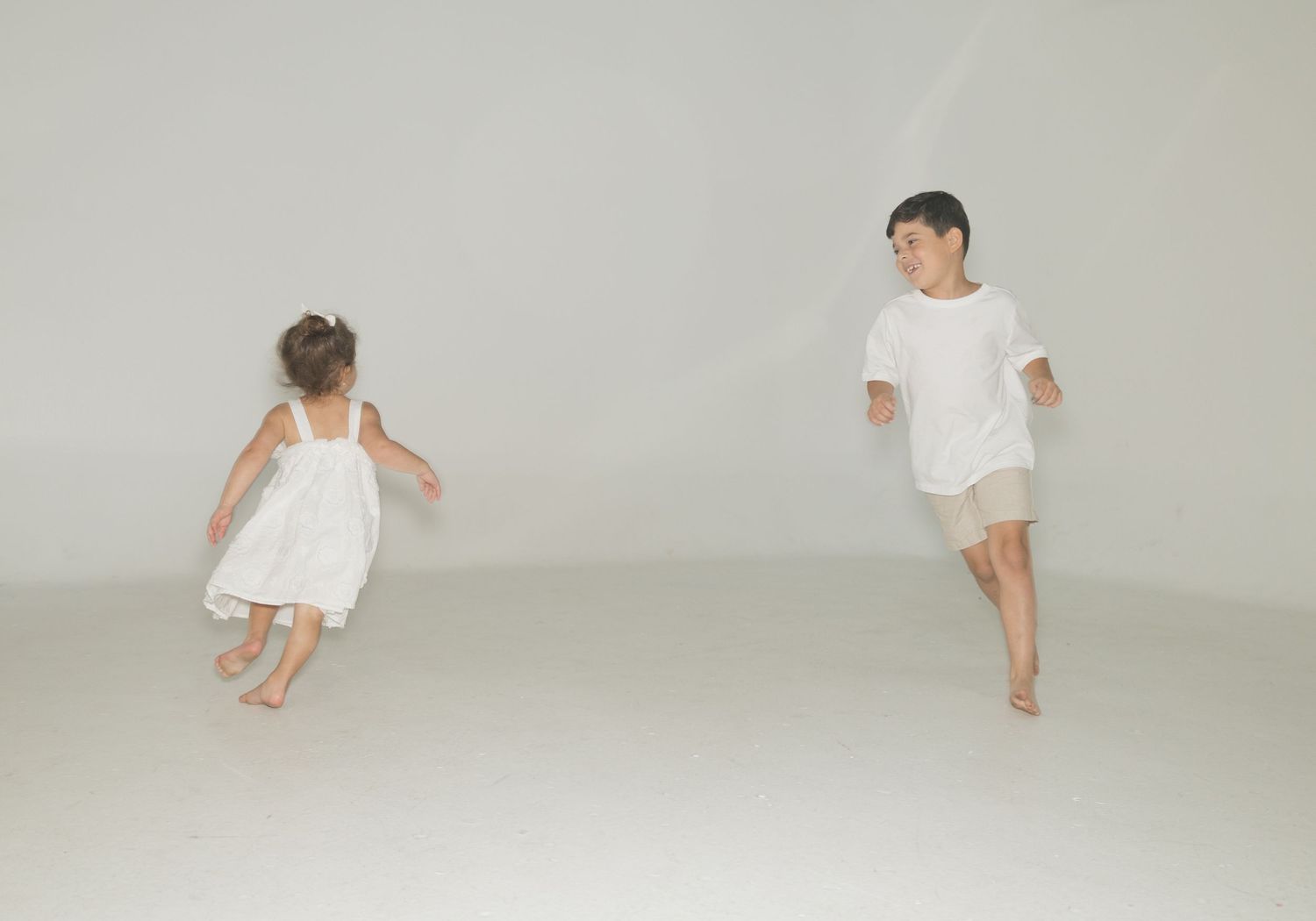 Two young children in white clothing playing and running barefoot in a bright white open space.