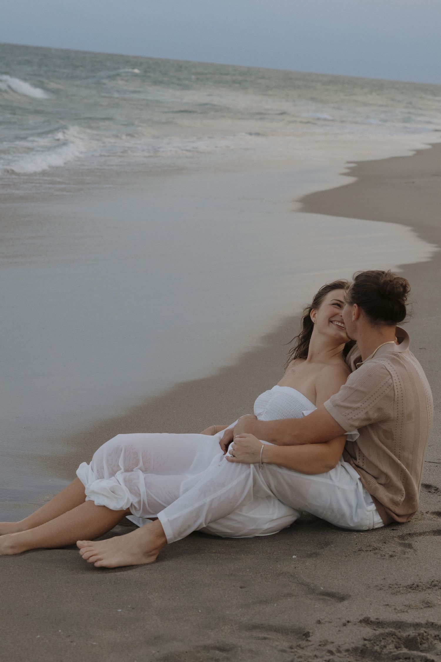Couple dressed in white relaxing together on a sandy beach at sunset creates romantic coastal mood.