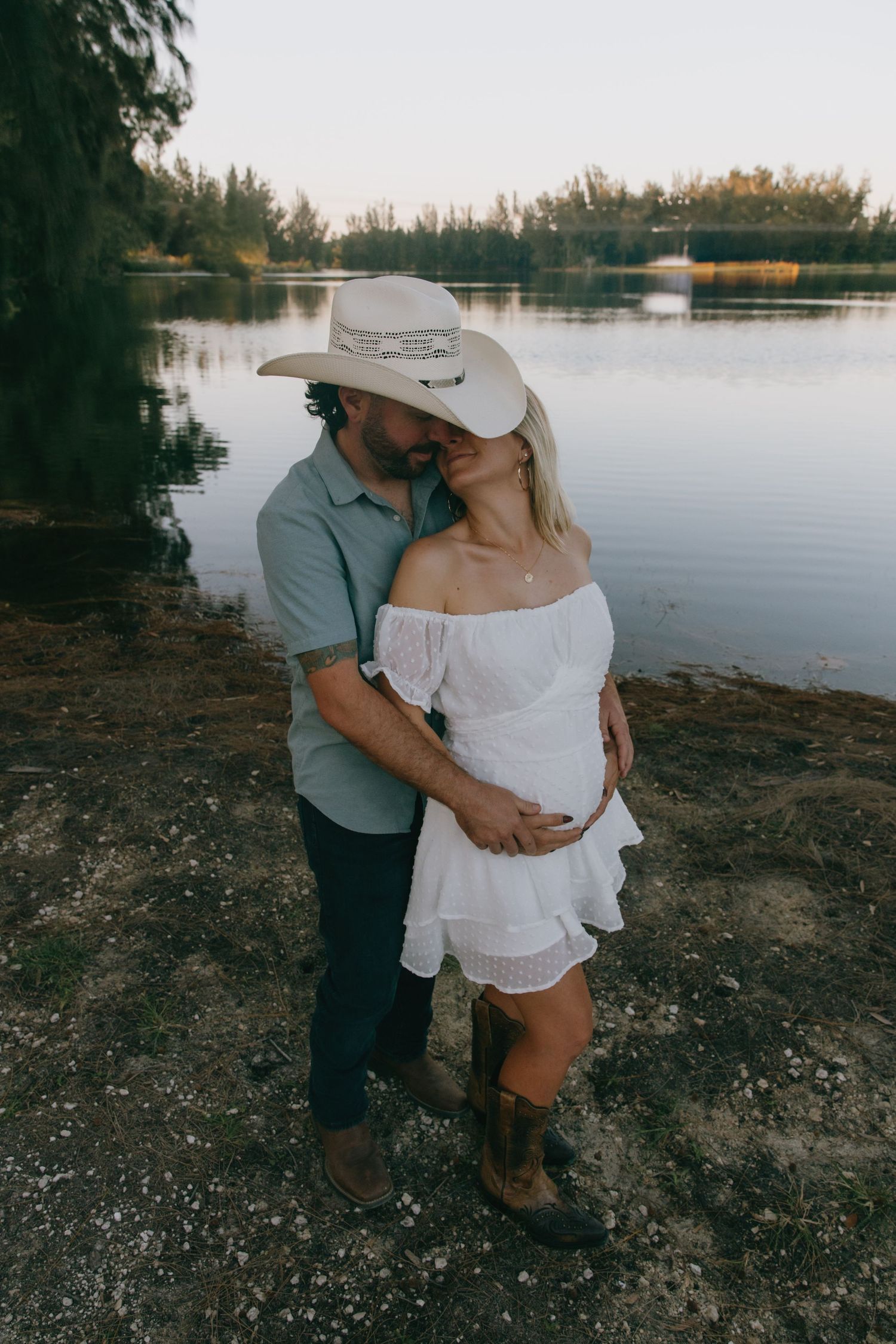 A couple in western wear shares an intimate moment by a peaceful lake.