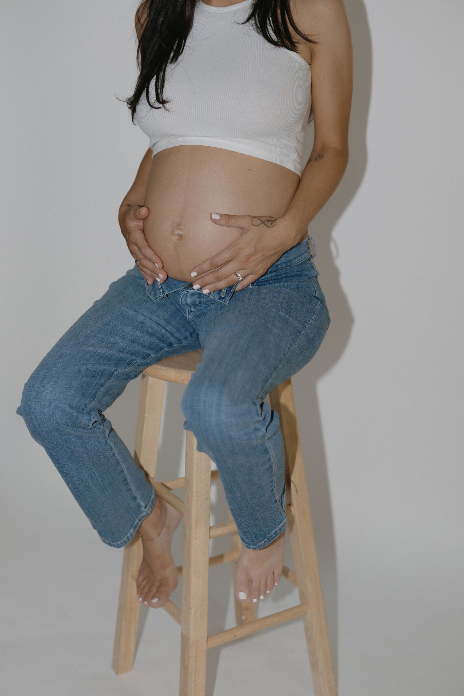 Person sitting on wooden stool wearing jeans and white top against white background.
