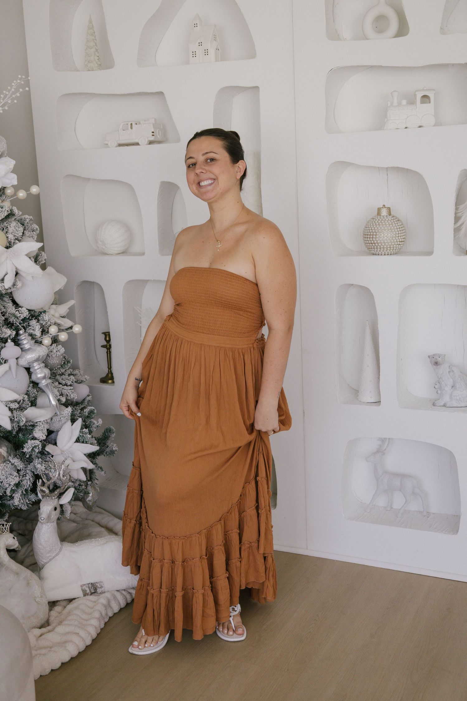 A person wearing a brown strapless maxi dress with ruffle details stands in front of a white decorative wall with built-in shelves.
