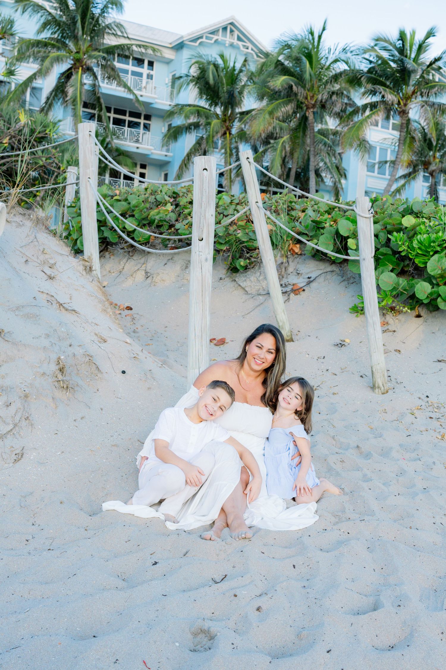 A beachside family portrait with palm trees and a coastal building in the background on a sandy beach.