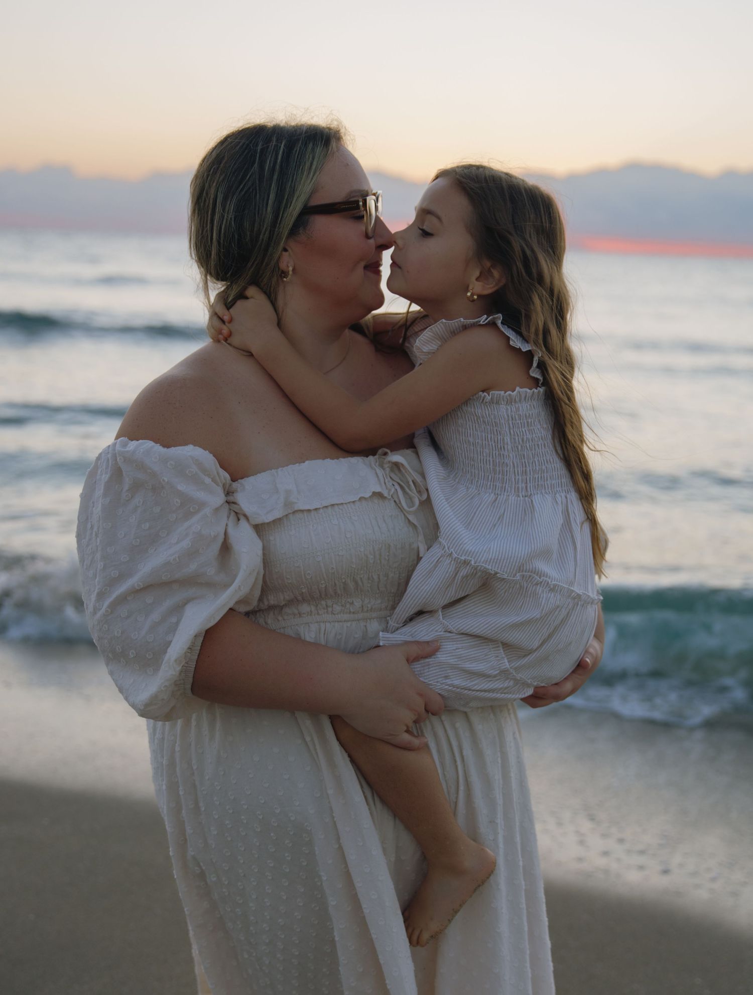 Two people in white dresses share an affectionate embrace on a beach at sunset.