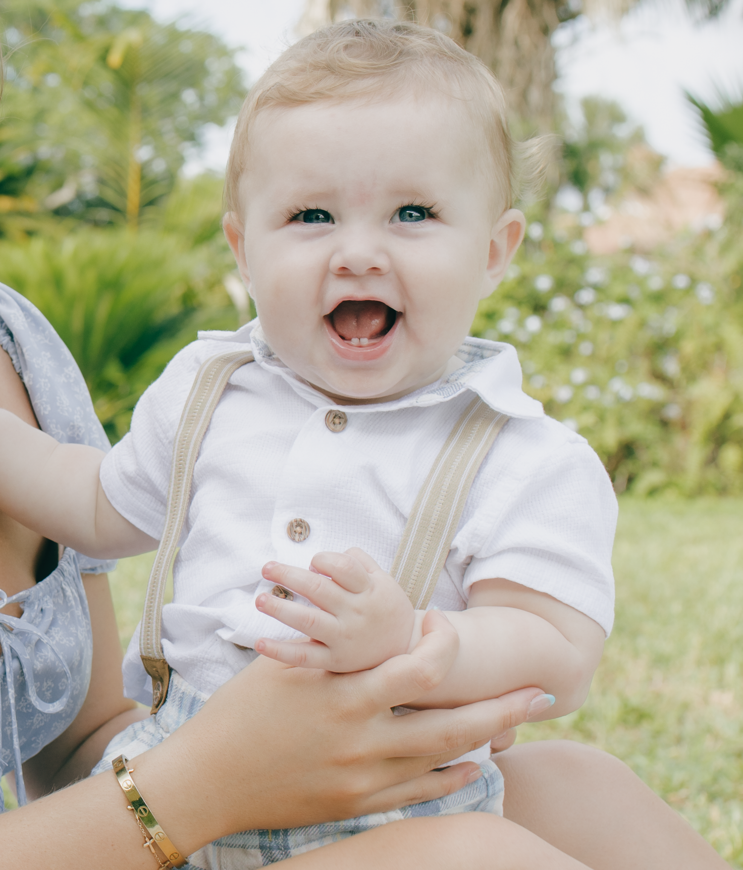 Adorable baby in white outfit and suspenders smiles and laughs while being held outdoors.