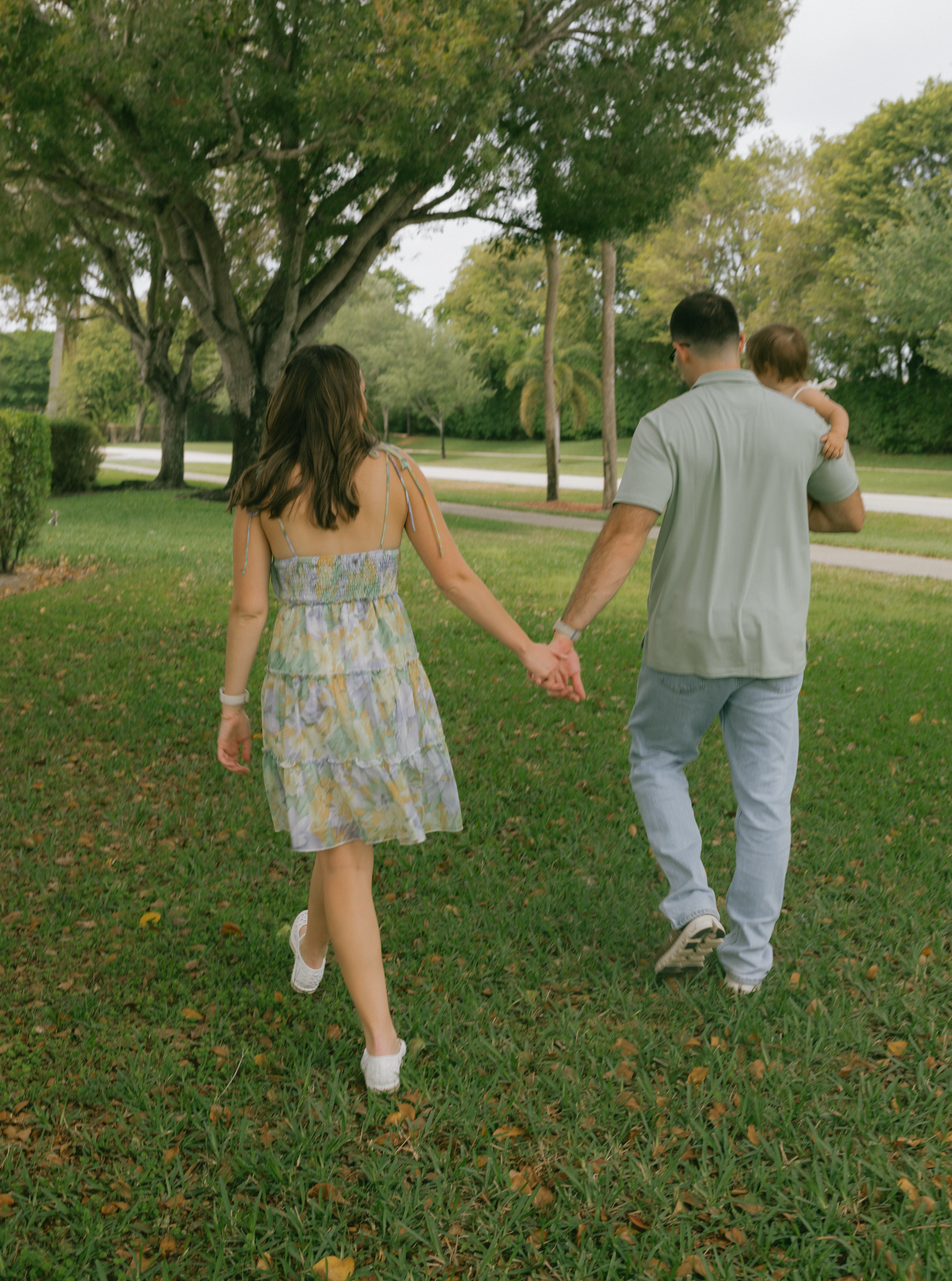 Two people walk hand in hand through a park with fallen autumn leaves scattered on the grass.