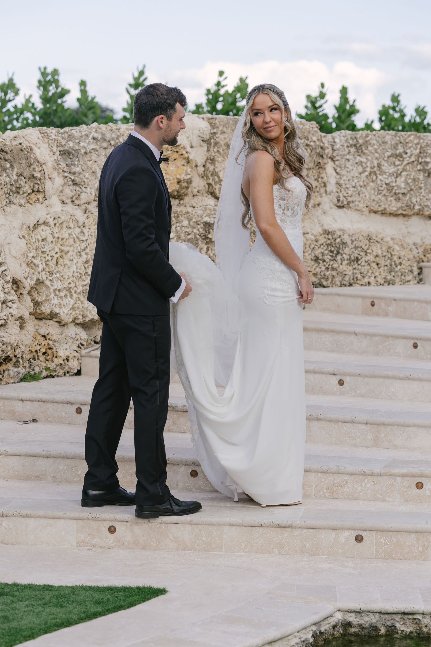 A bride in a flowing white dress and groom in black tuxedo stand on stone steps at an outdoor wedding venue.