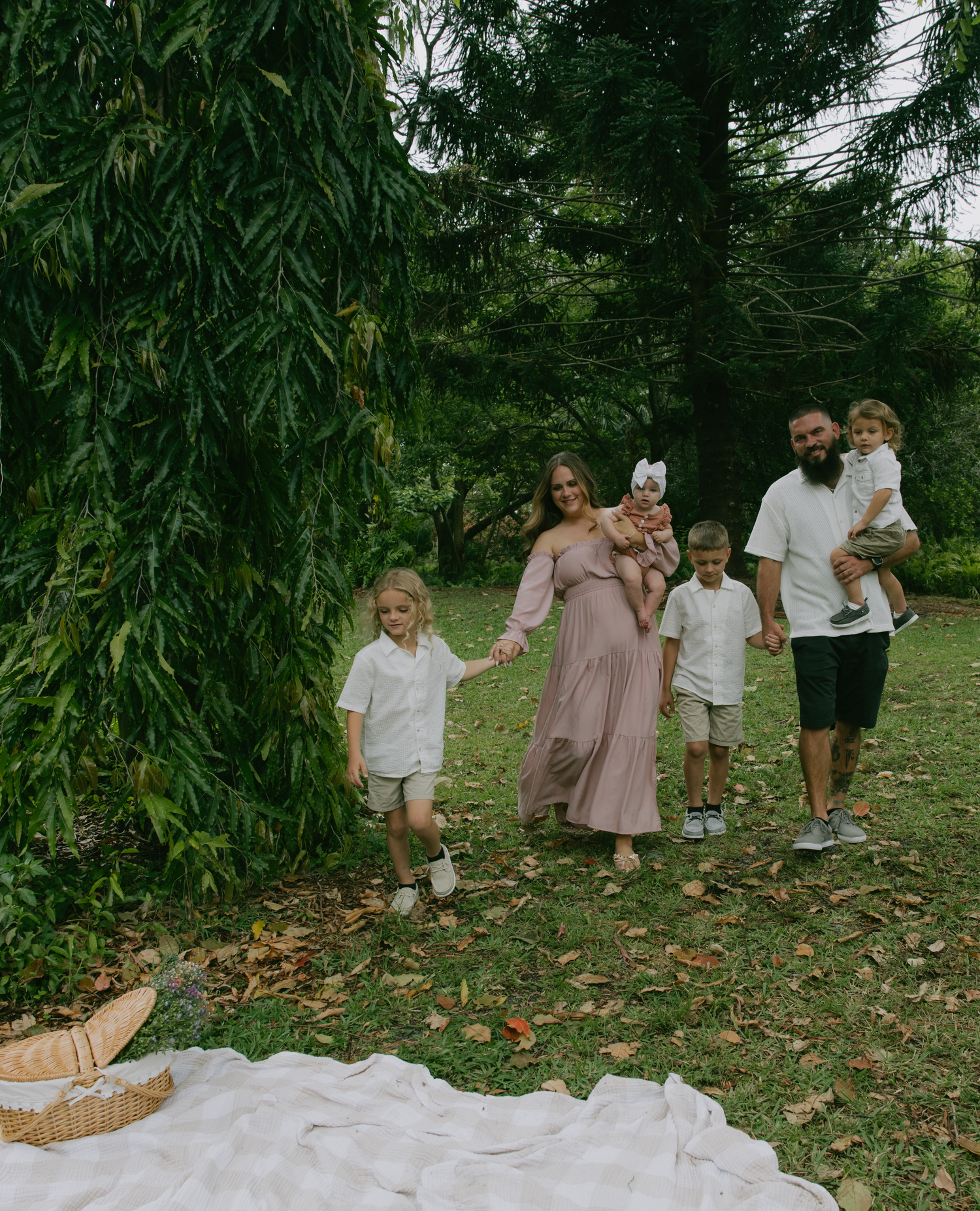 A group walks through a lush garden setting with a white blanket spread on the grass beneath tall willow trees.