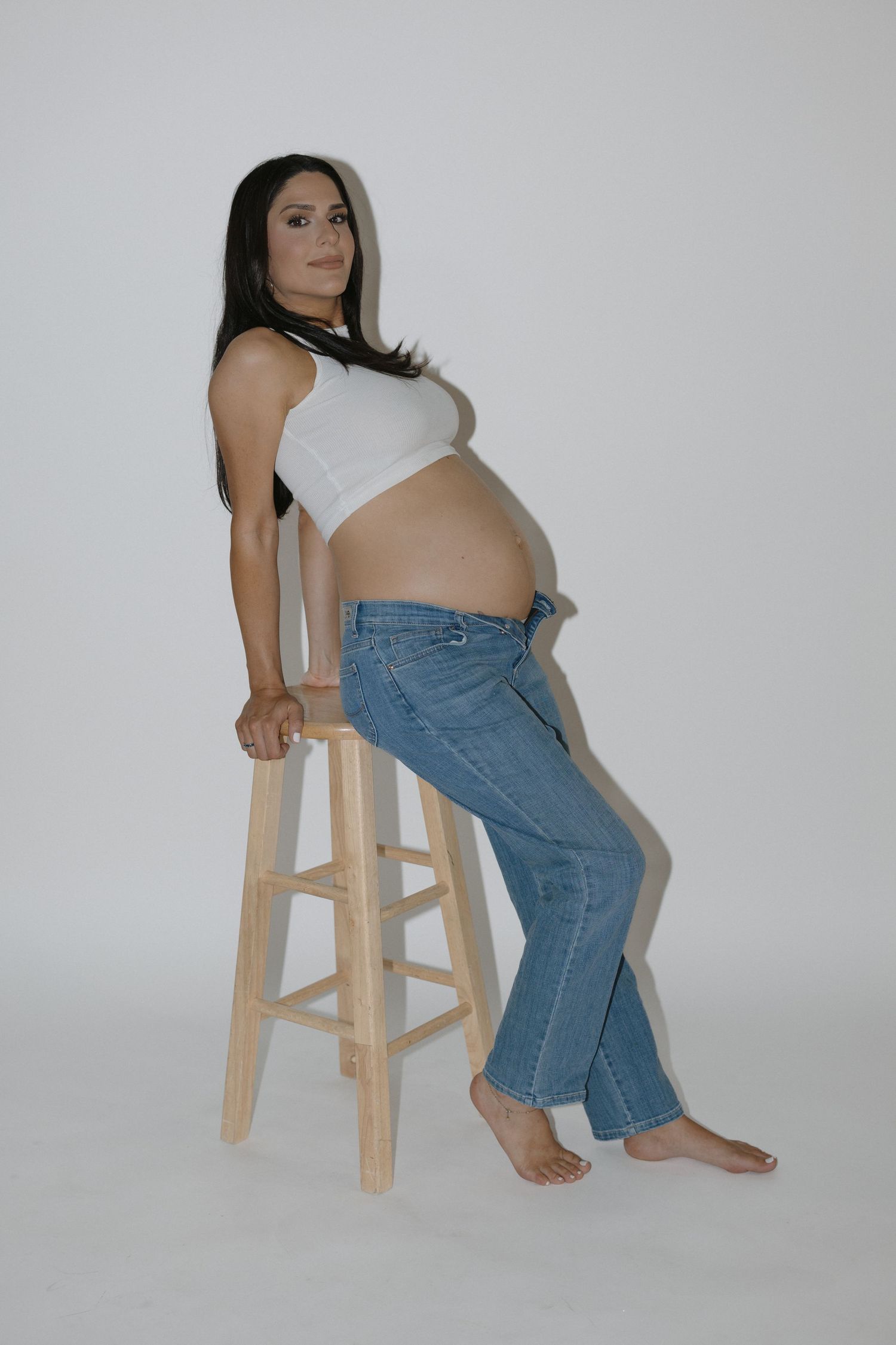 Artistic pregnancy photo series featuring poses on wooden stool in jeans and white top.
