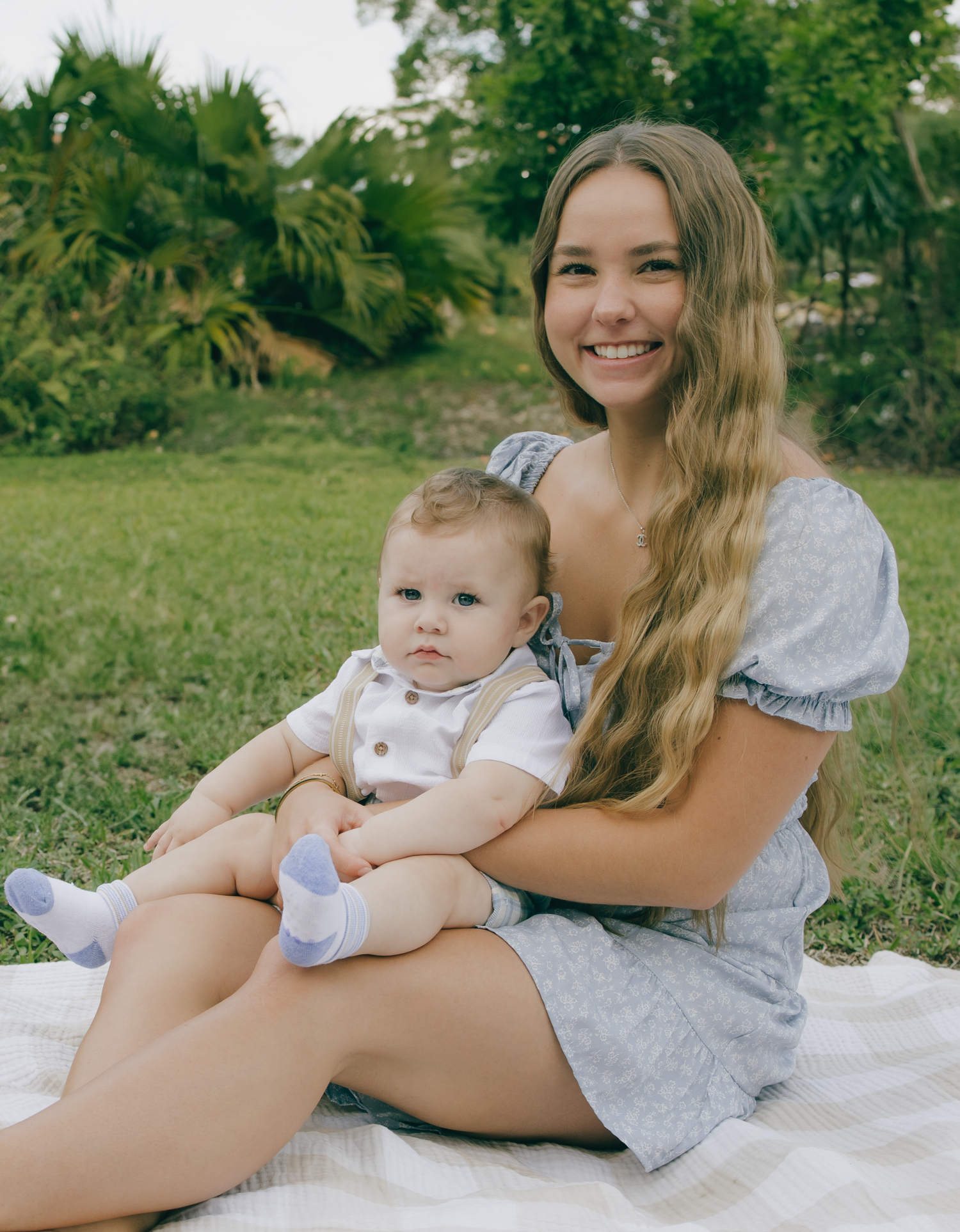 A cheerful outdoor portrait of a mother and baby sitting together on a white blanket.