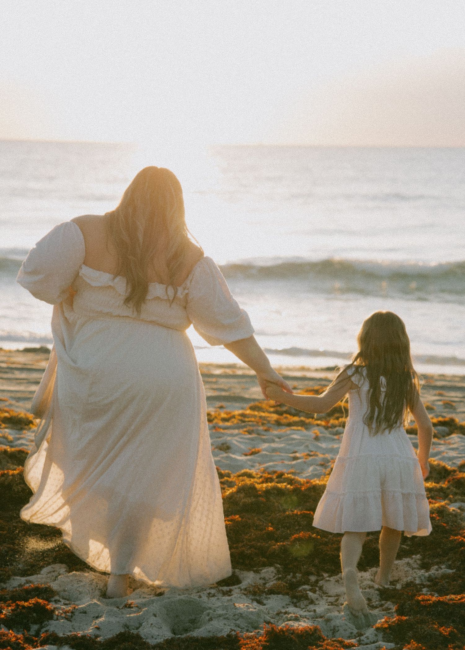 People in white dresses walking along golden sand beach during sunset.