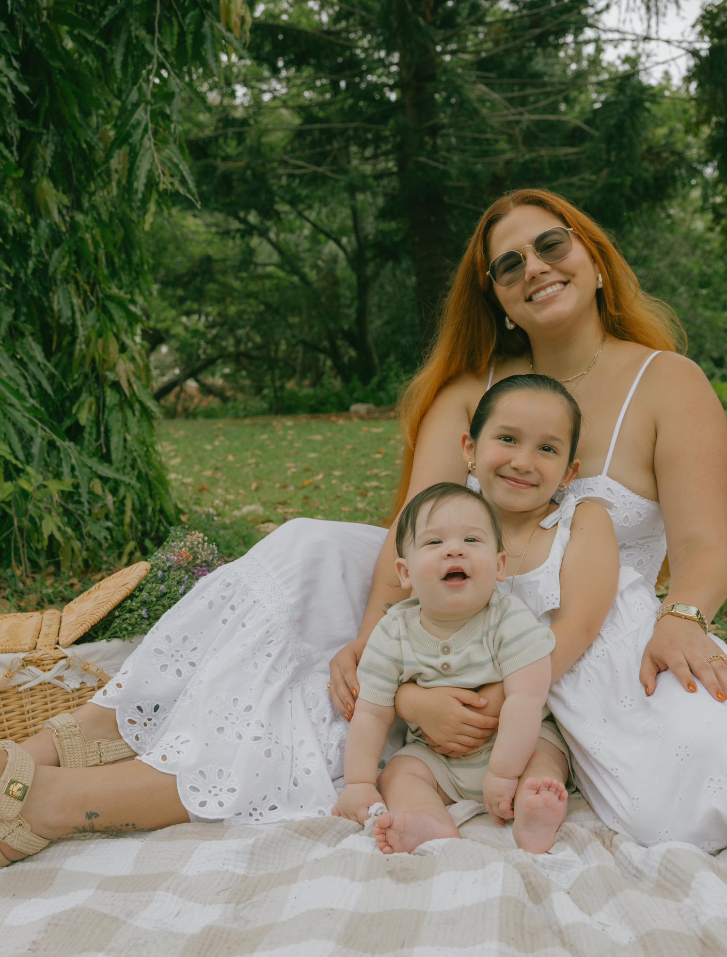 Woman and children sitting on picnic blanket outdoors surrounded by lush greenery.