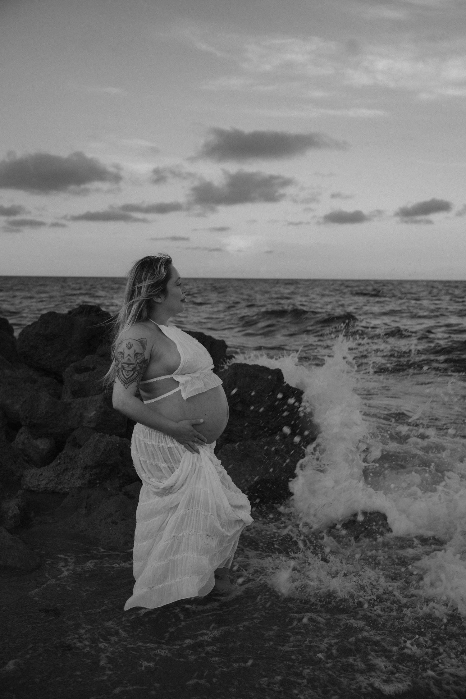 Black and white maternity photo of woman on rocky coastline with crashing waves.