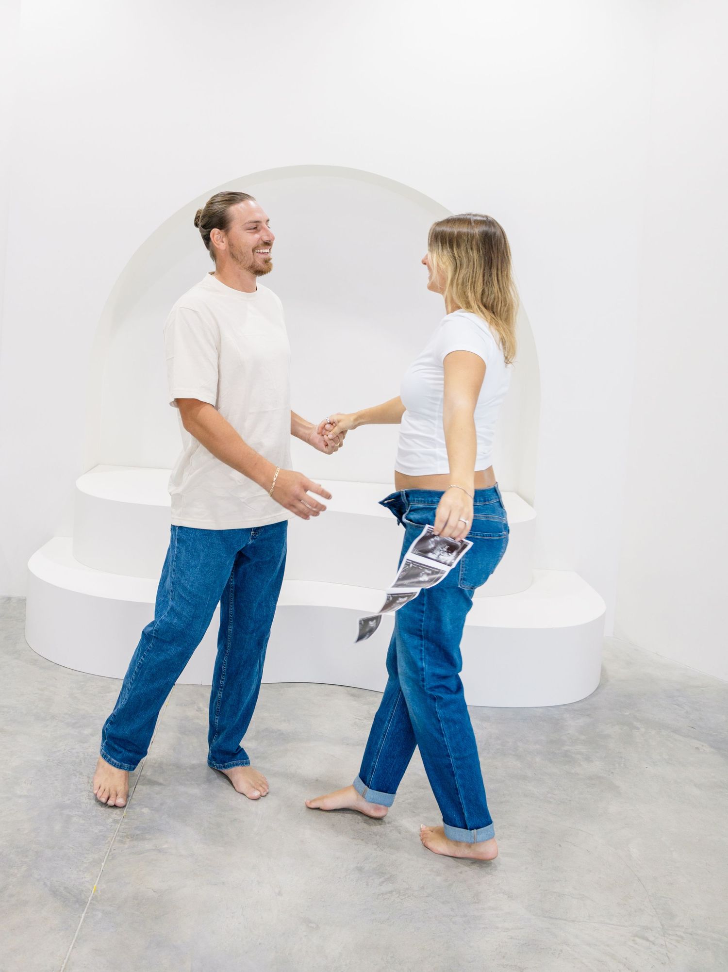 A couple in matching blue jeans and white tops shake hands and smile in a modern white gallery space.