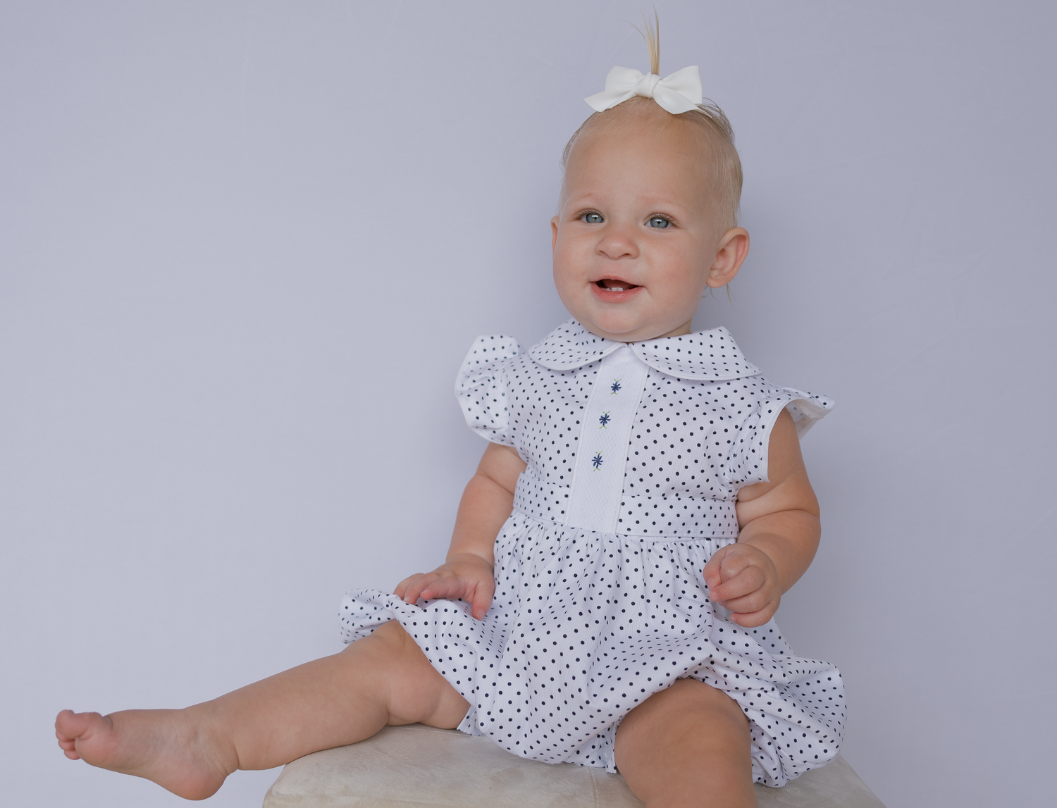 Baby wearing white polka dot romper sits and plays on light gray backdrop in multiple poses.