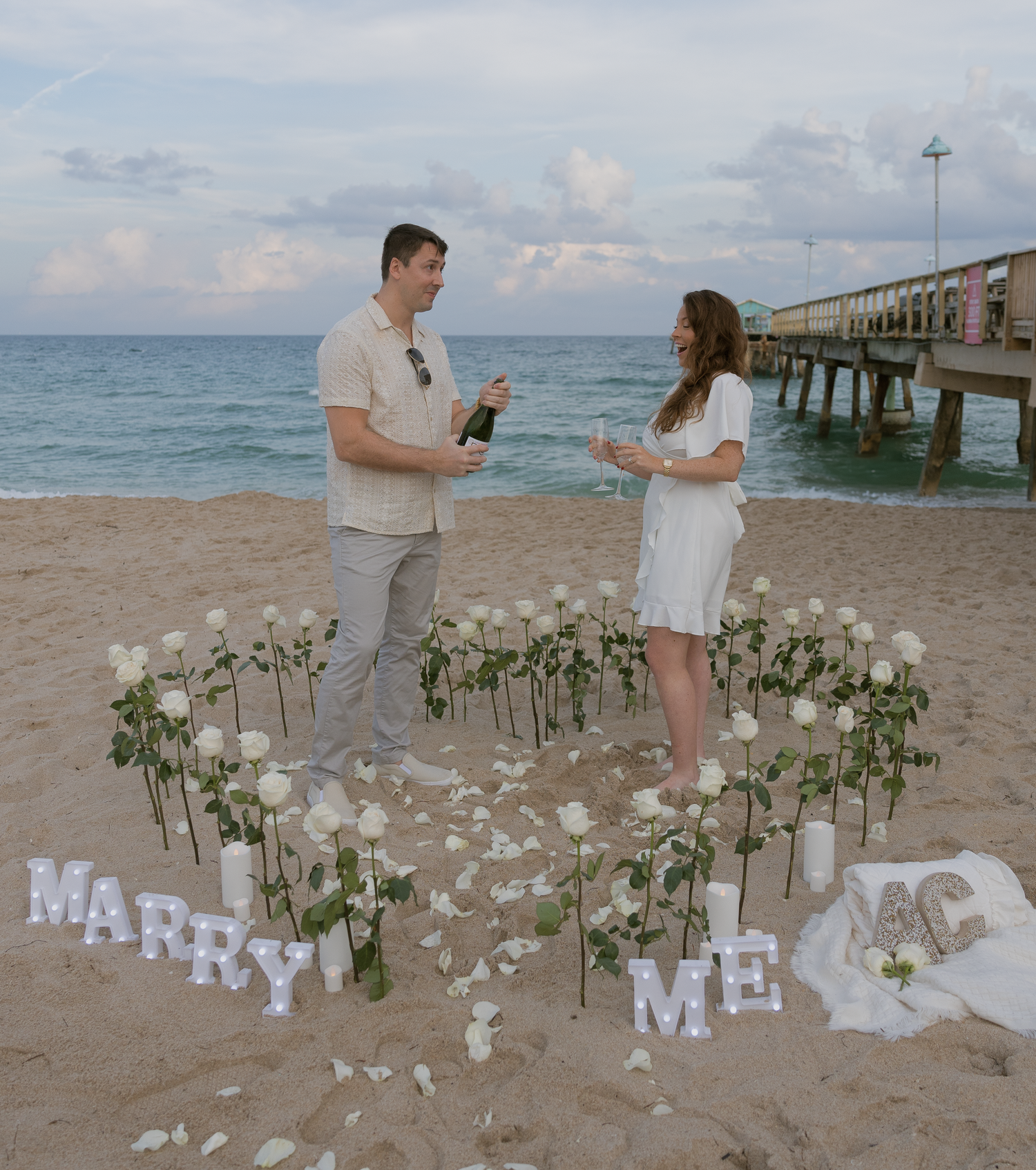 A romantic beach proposal setup with white flowers arranged in a heart shape and 'MARRY ME' letters on the sand at sunset.