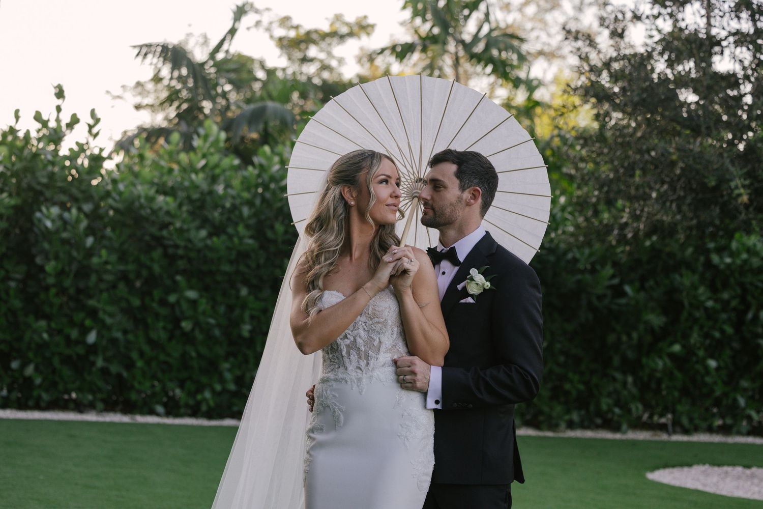 A couple poses together under white parasols in a lush garden setting during their wedding celebration.