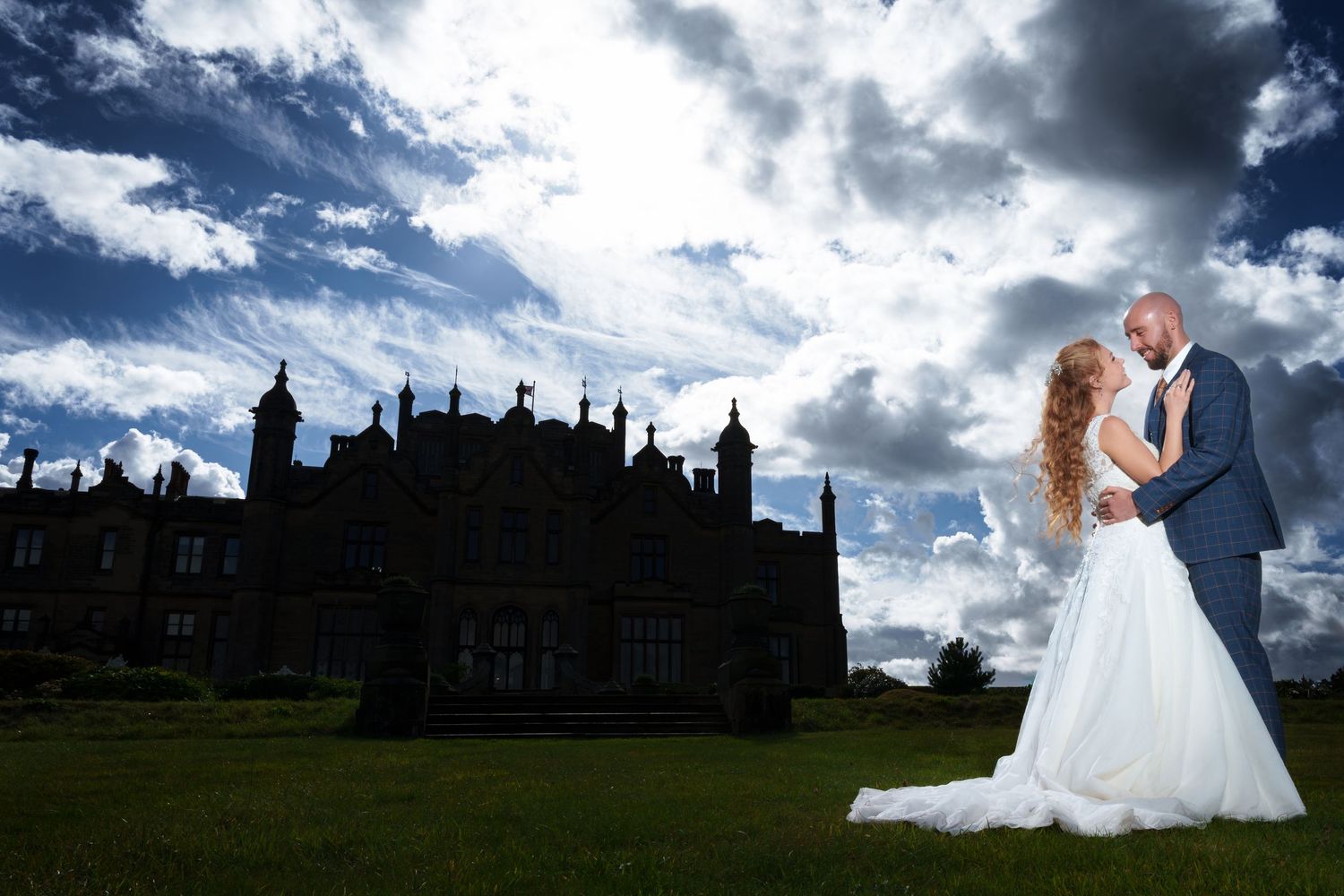 A couple in elegant wedding attire poses against dramatic cloudy skies near a historic estate building.