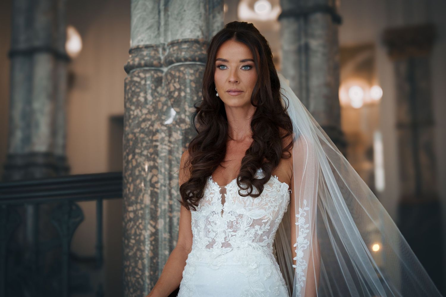 A bride in a lace gown poses elegantly in front of a weathered stone column with soft lighting in the background.