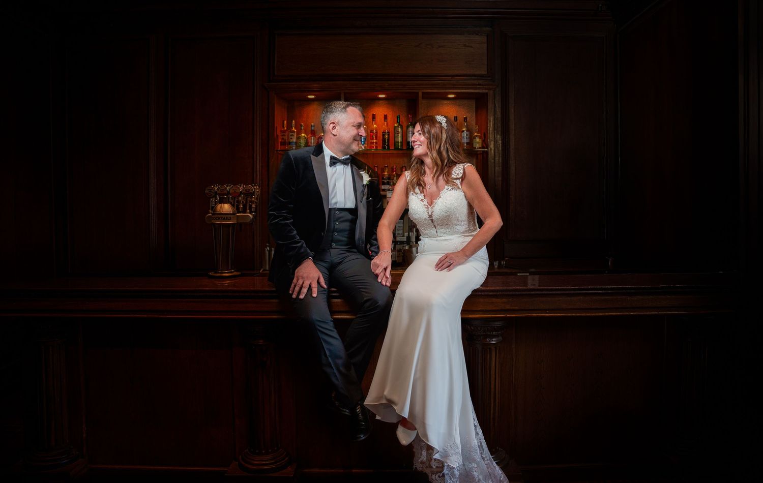 Elegant bride and groom in wedding attire holding hands, smiling at each other in a dark wood-paneled venue.