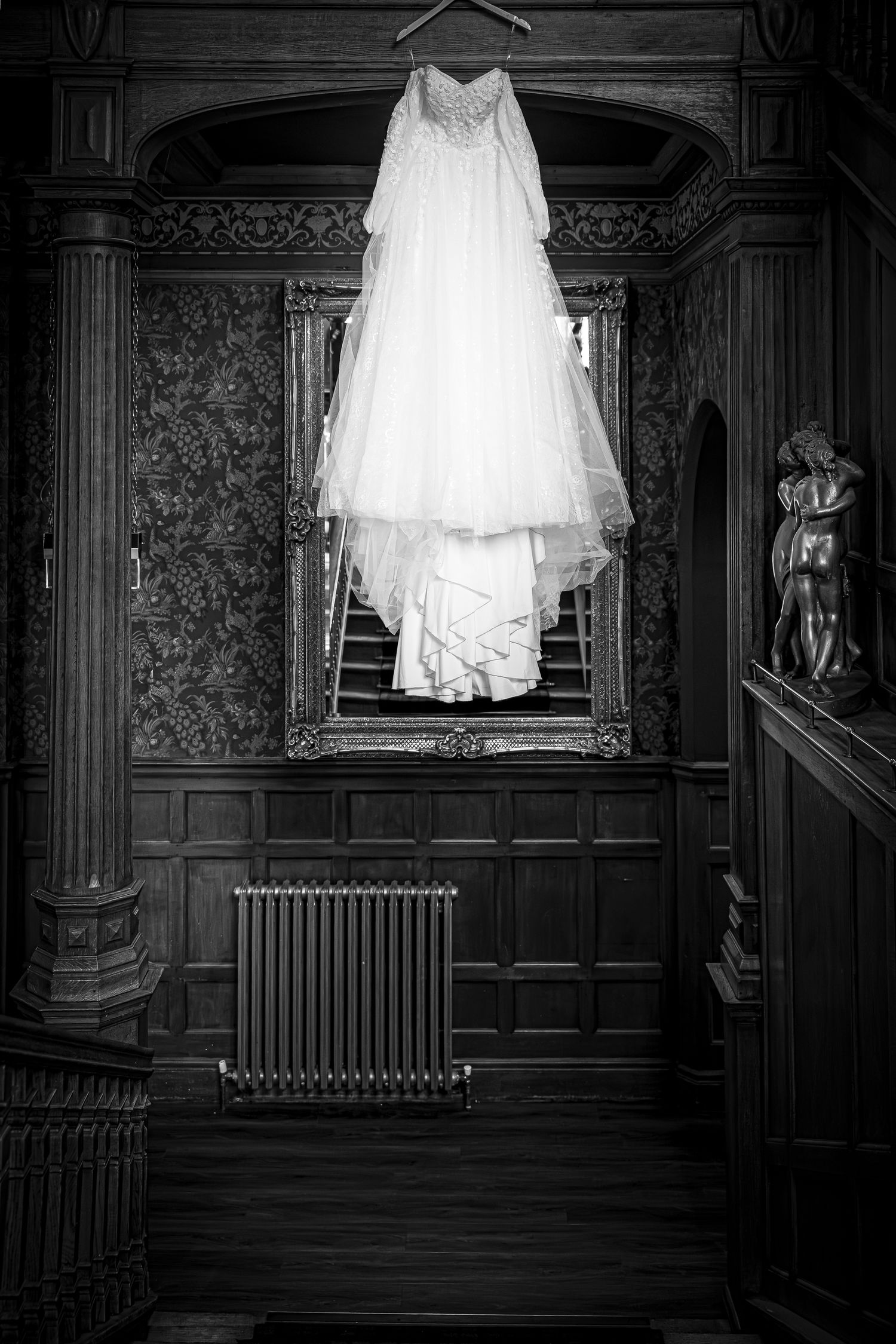 Black and white photo of a white wedding dress hanging in an ornate Victorian interior with wood paneling and decorative mirror.