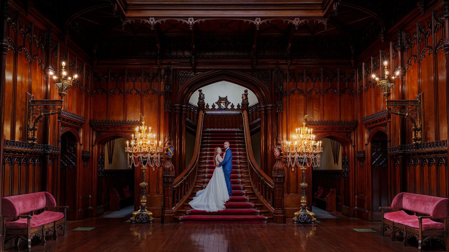 An elegant wood-paneled grand staircase hall with crystal chandeliers and pink velvet benches in a historic mansion interior.
