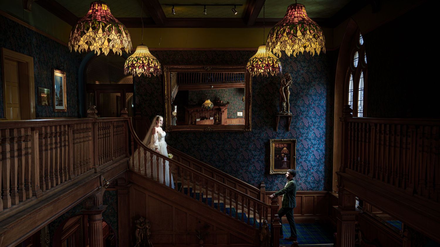 Bride in white gown stands on ornate wooden staircase under Tiffany chandeliers in elegant Victorian mansion.