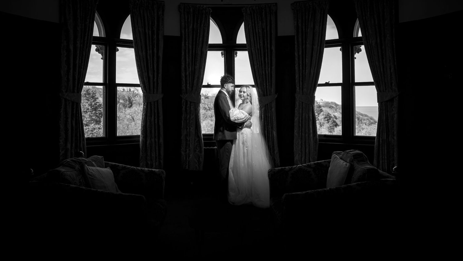 Black and white photo of a bride and groom embracing silhouetted against tall arched windows in an elegant room.