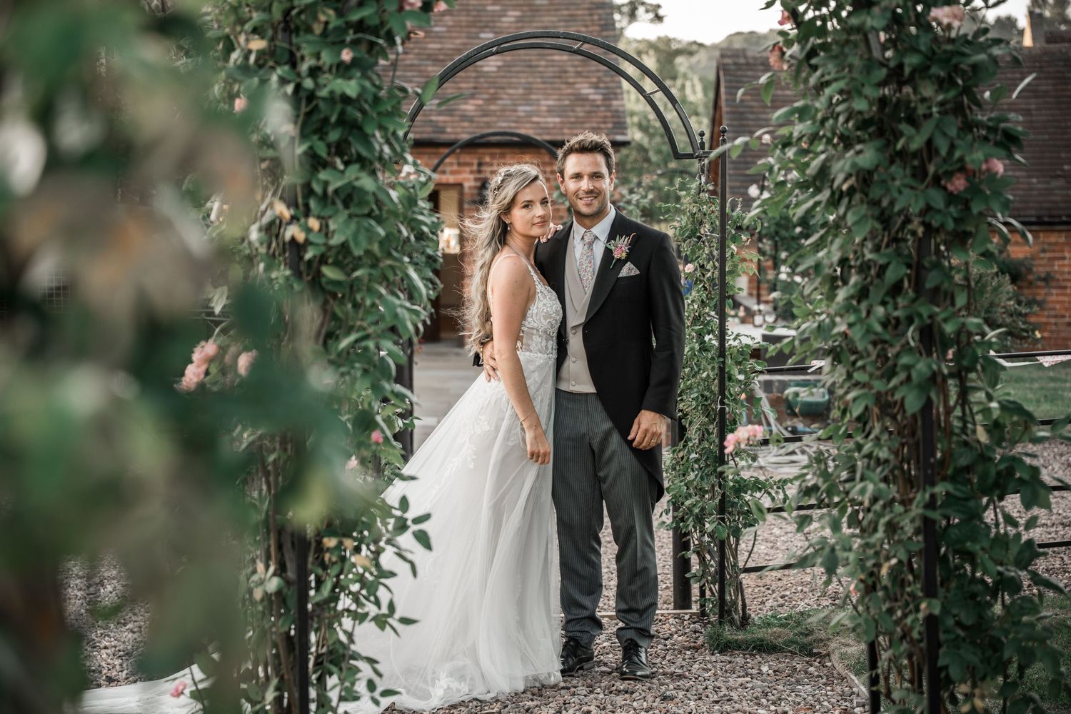 A couple in wedding attire stands beneath a romantic garden archway covered in climbing vines and greenery.