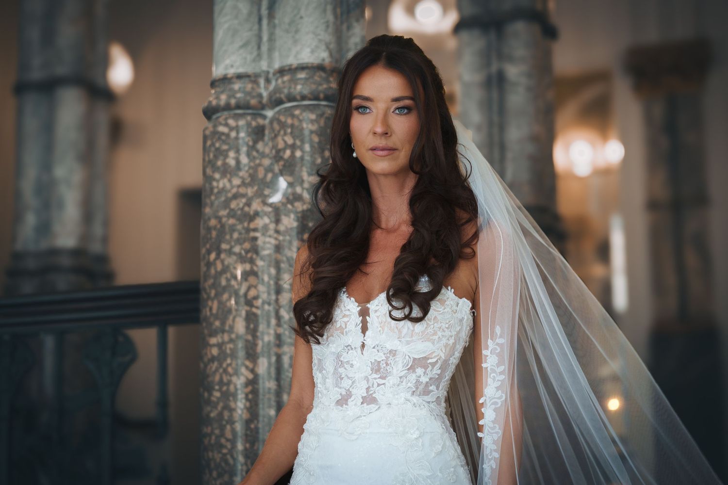 A stunning bride in a white lace wedding dress poses elegantly inside an ornate stone building with romantic lighting.