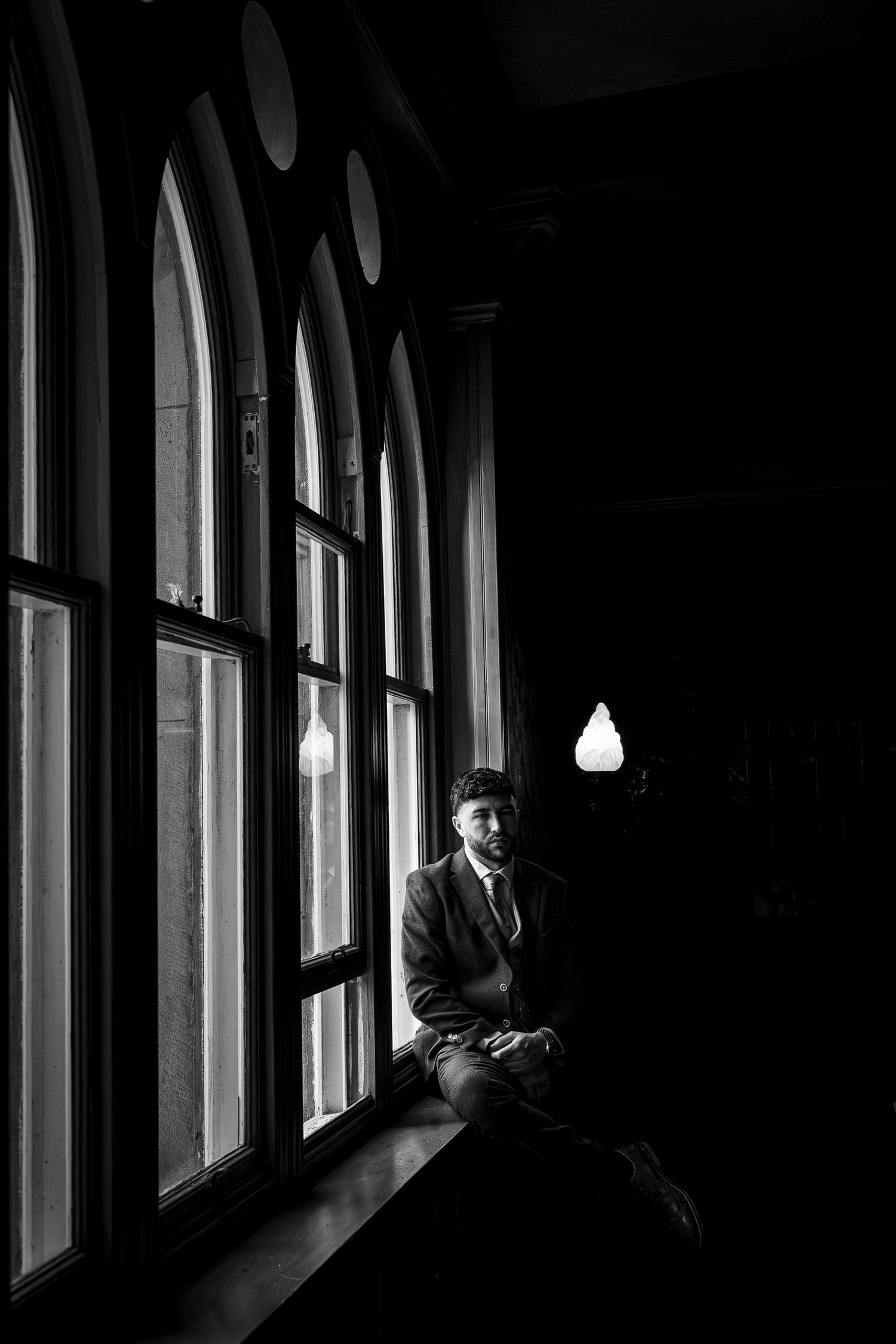 Black and white photo of a man in a suit sitting on a windowsill in a dark, gothic-style building.