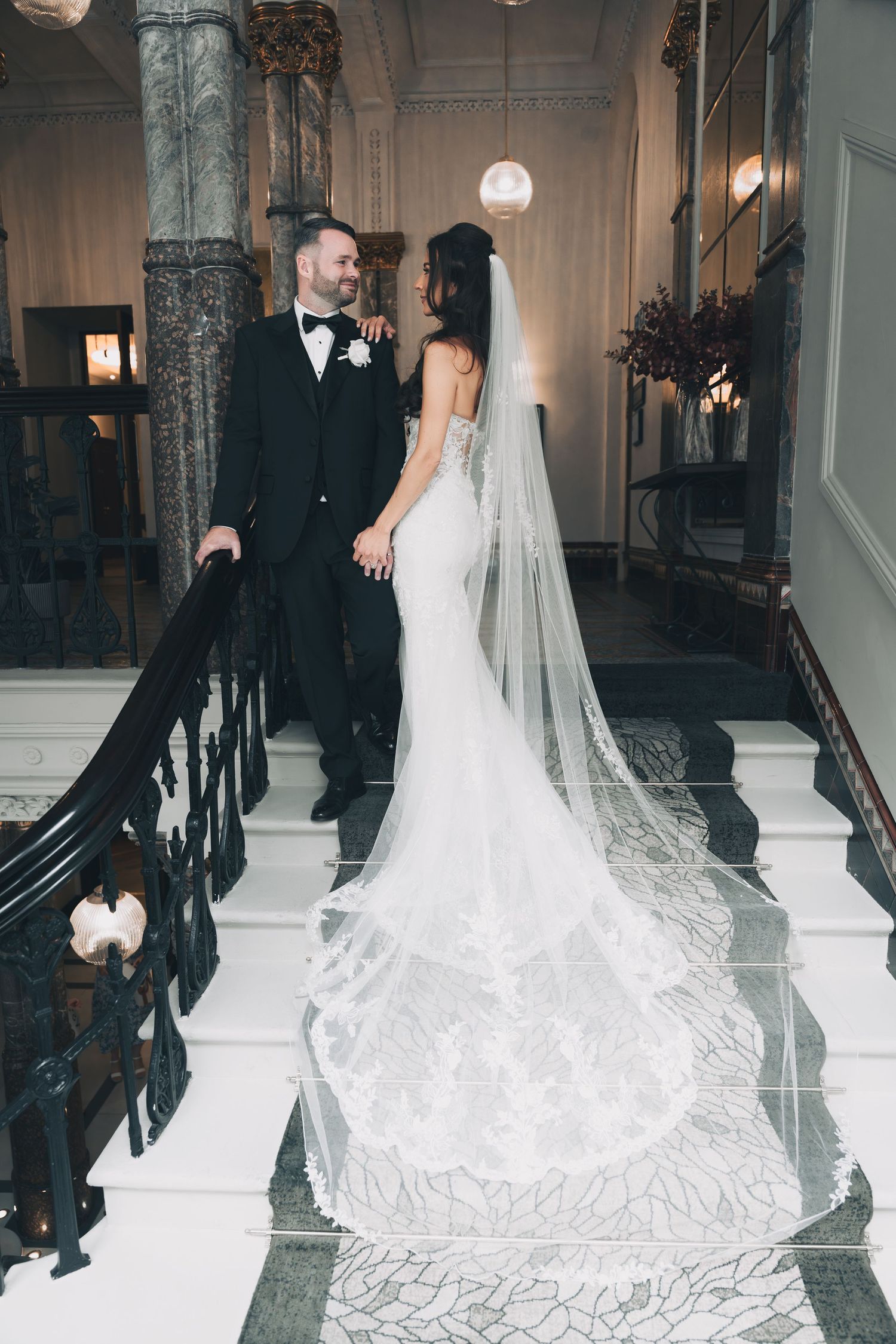 A bride and groom walk together down an elegant hallway with marble columns and sparkling chandeliers overhead.