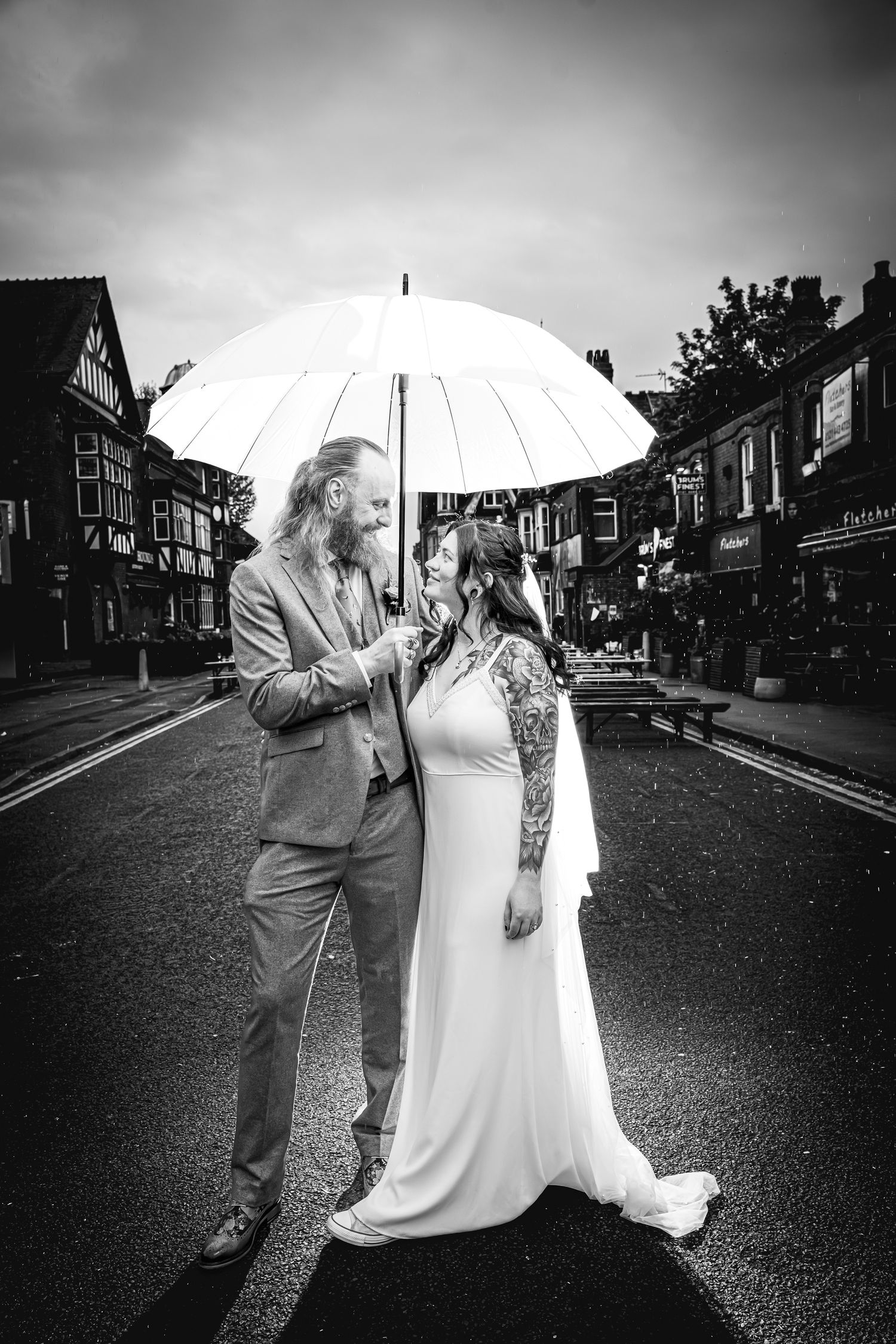 Black and white photo of a bride and groom sharing a white umbrella on a street, smiling at each other on their wedding day.
