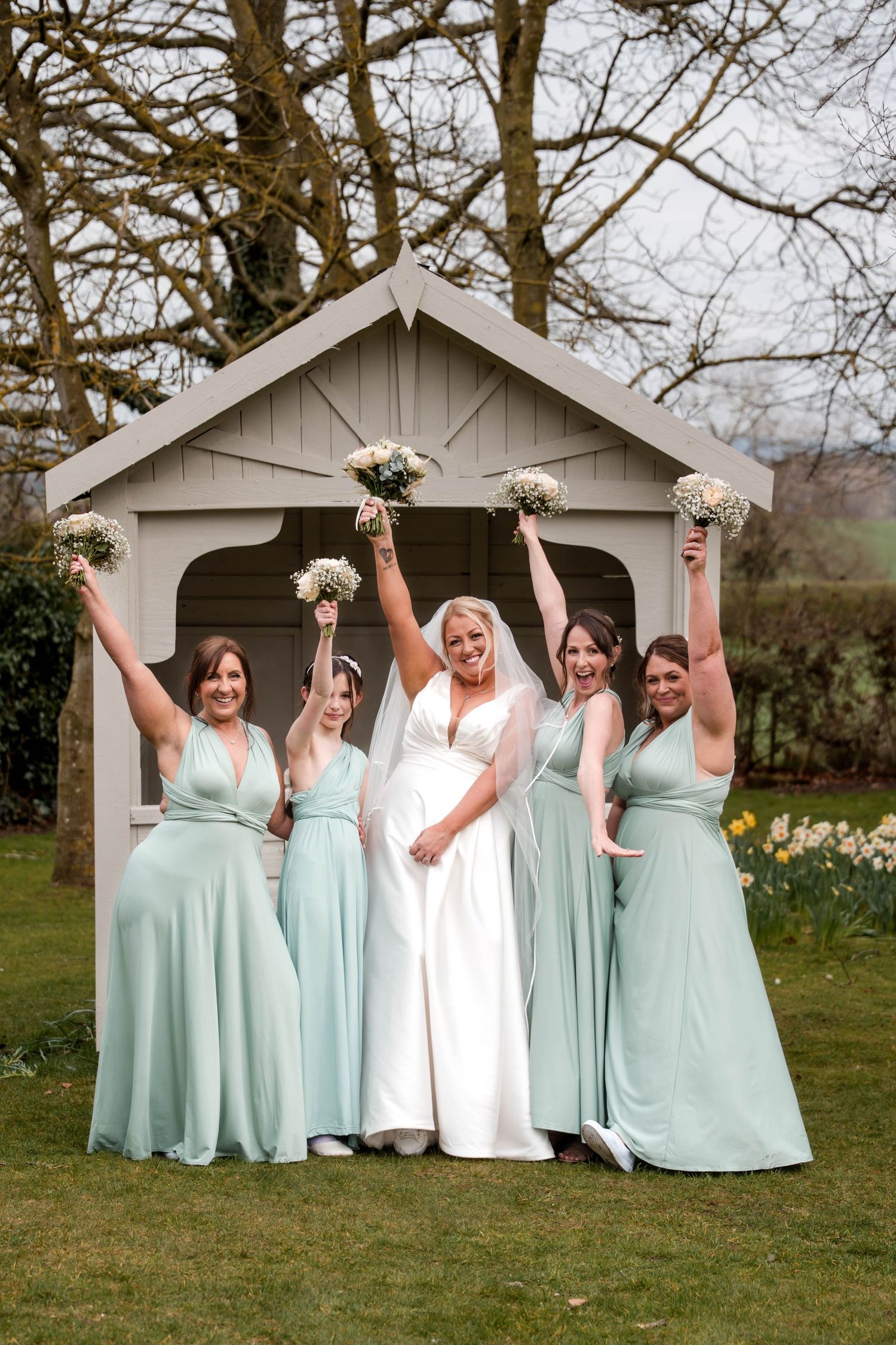 Bridesmaids in mint green dresses pose joyfully with bride outside white garden pavilion.