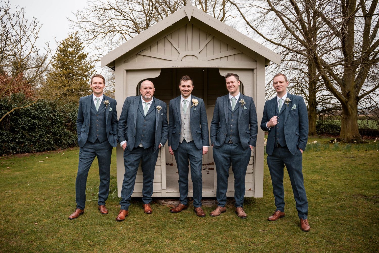 Groomsmen in navy suits lined up posing together in front of a white wooden building.