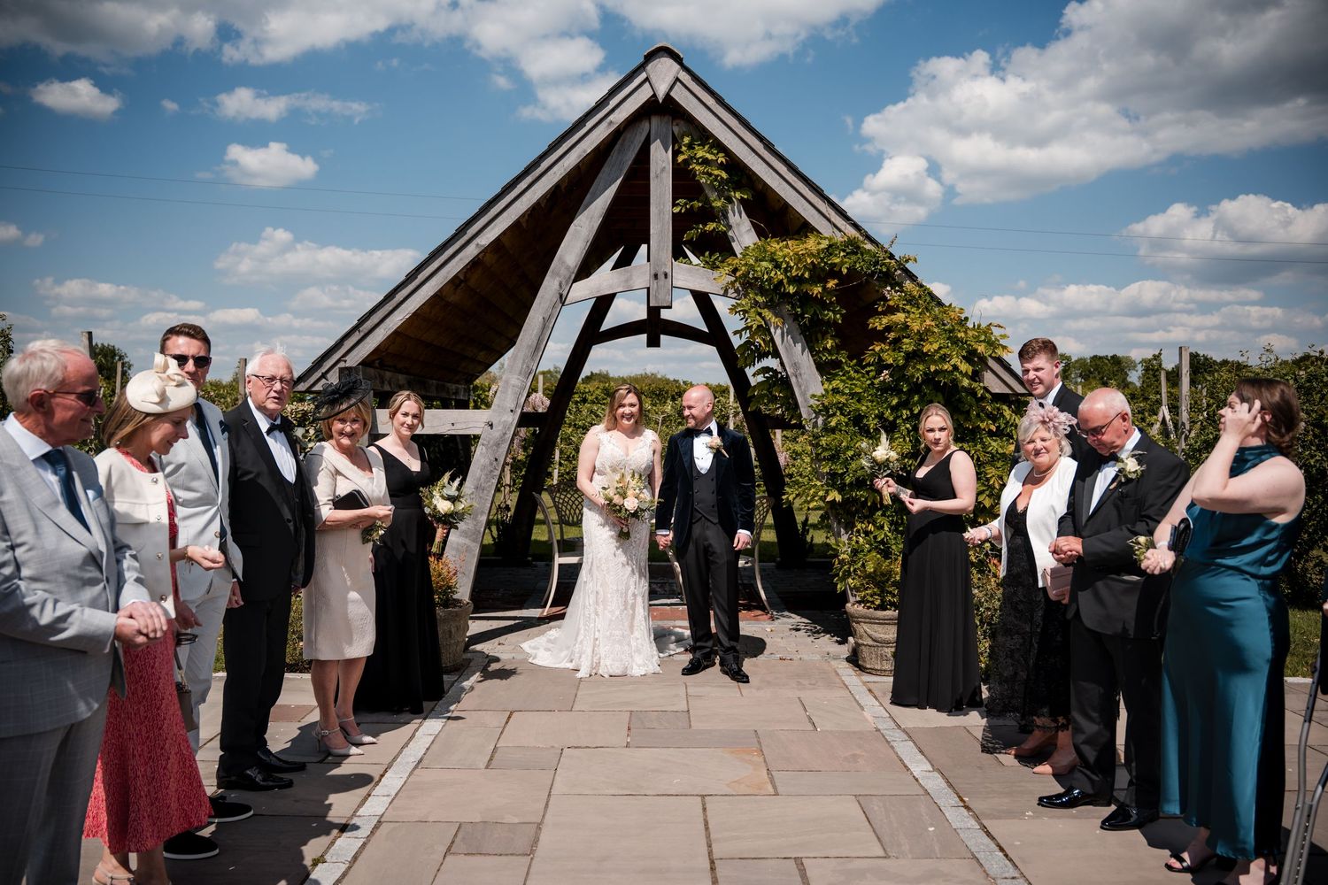 Wedding ceremony guests line the aisle under a wooden arch structure on a sunny day.