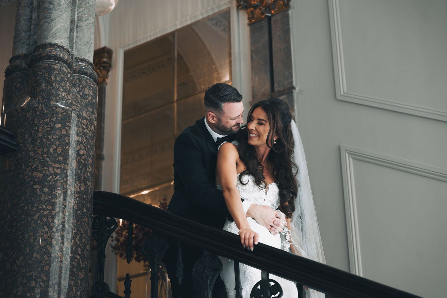 A black and white wedding photo shows a couple sharing an intimate moment in an elegant venue hallway.