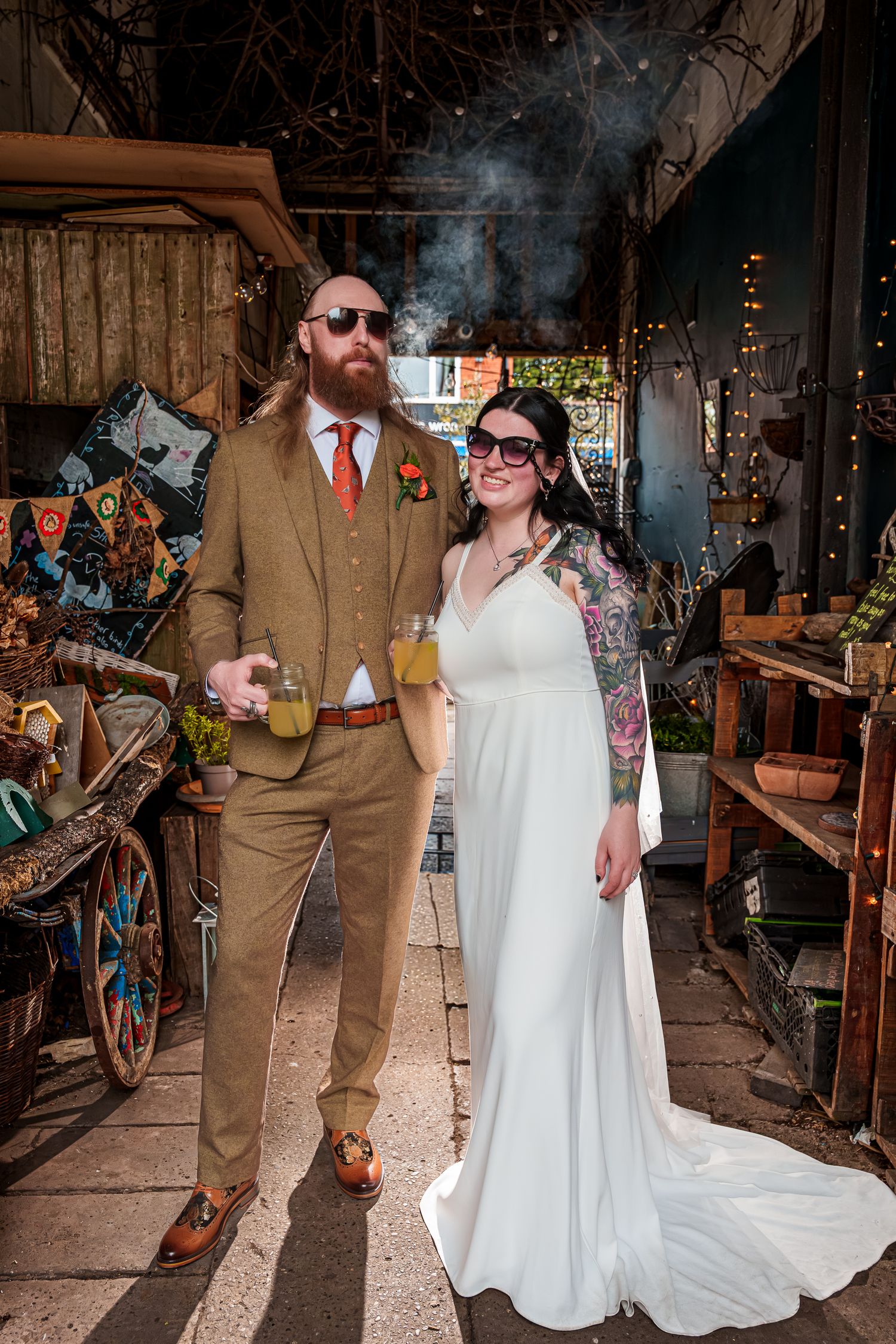 Tattooed bride in white gown and groom in tan suit with orange tie pose together at rustic wedding venue.