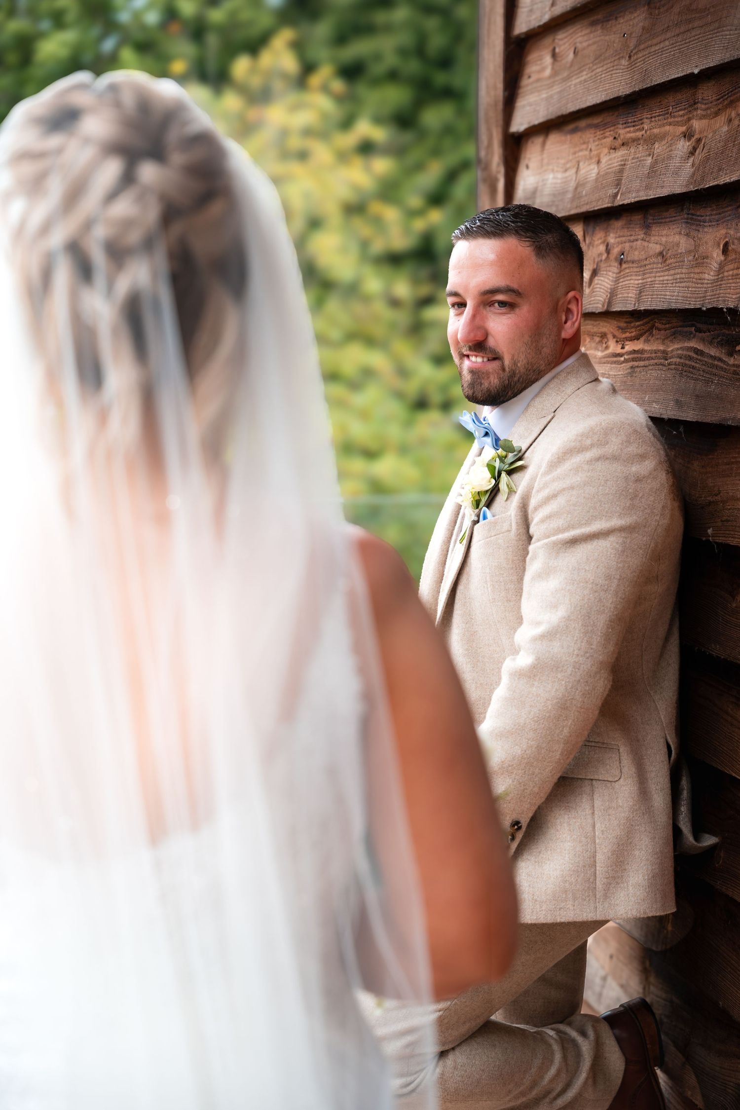 A groom in a beige suit leans against a rustic wooden wall during wedding photos.
The Mill Barns wedding venue