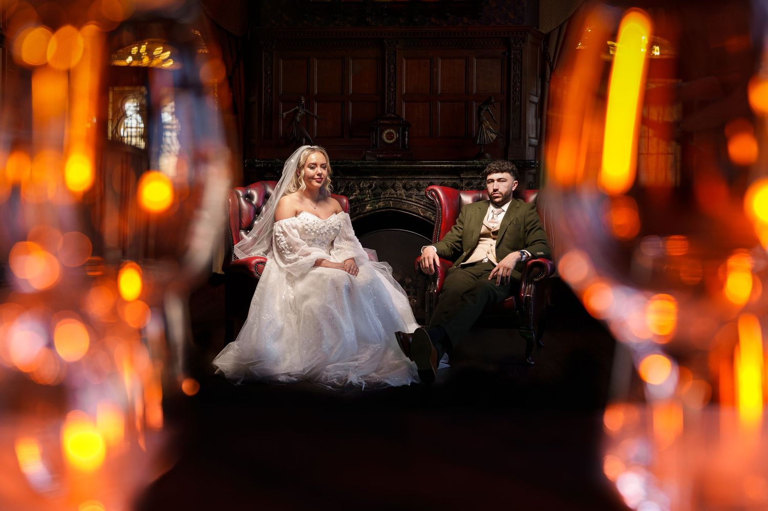 Bride and groom seated in ornate chairs framed by glowing candles in an elegant candlelit wedding venue.