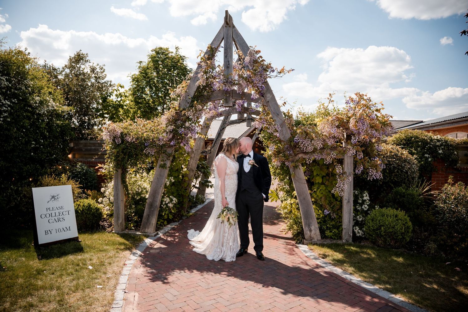 Wedding party gathered at garden venue under wooden arbor on bright summer day.