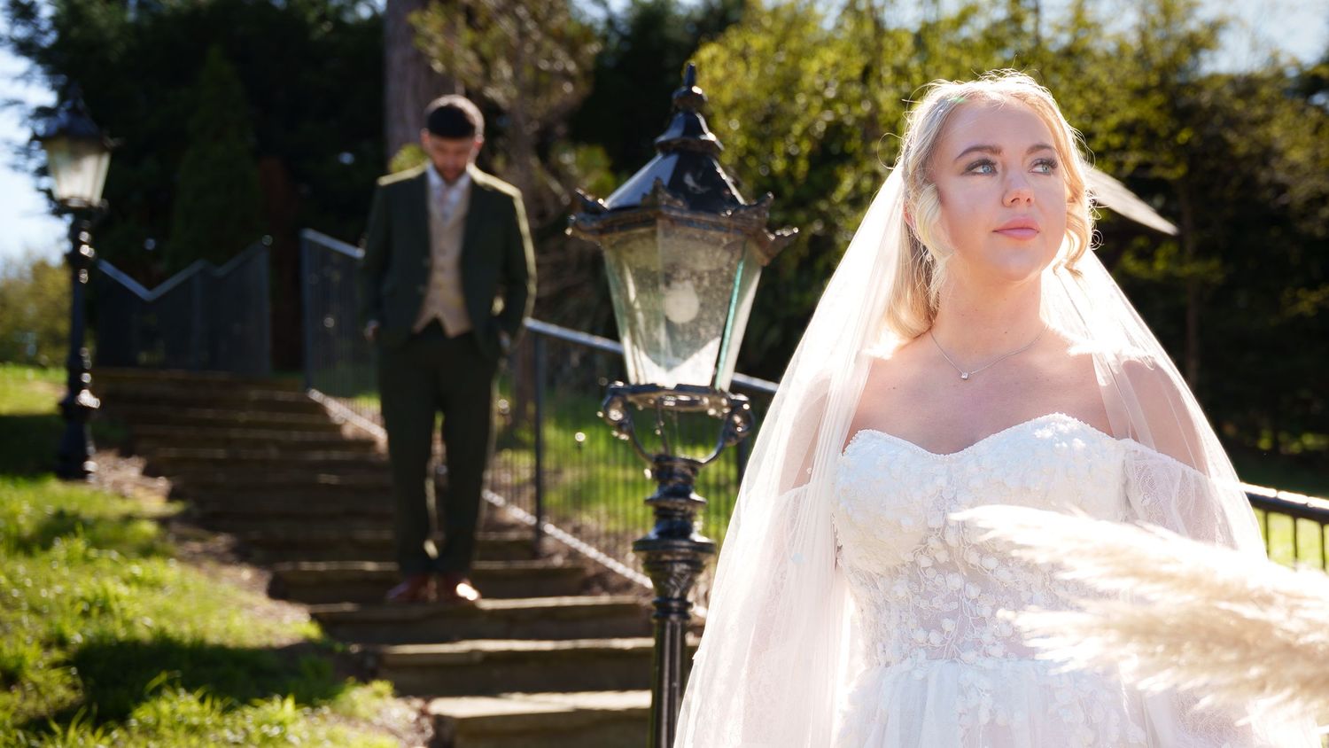 Bride in strapless lace wedding gown and veil stands outdoors while groom in suit descends stone steps behind her.