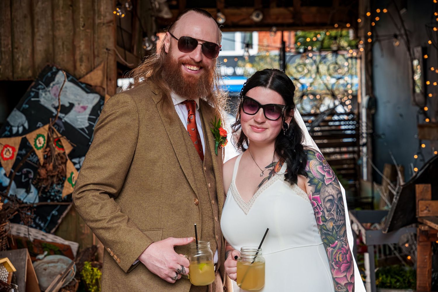 Tattooed bride in white dress and bearded groom in tweed suit holding cocktails at rustic wedding venue.