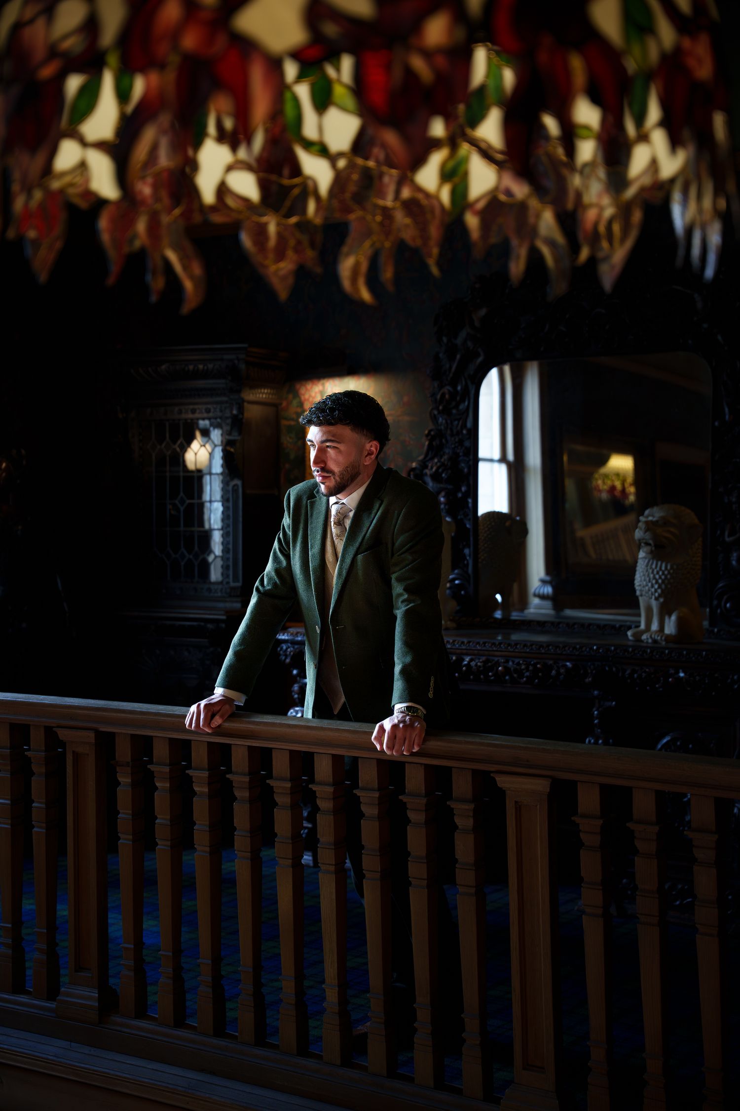 Groom in green suit standing at wooden railing beneath stained glass ceiling in dark interior.