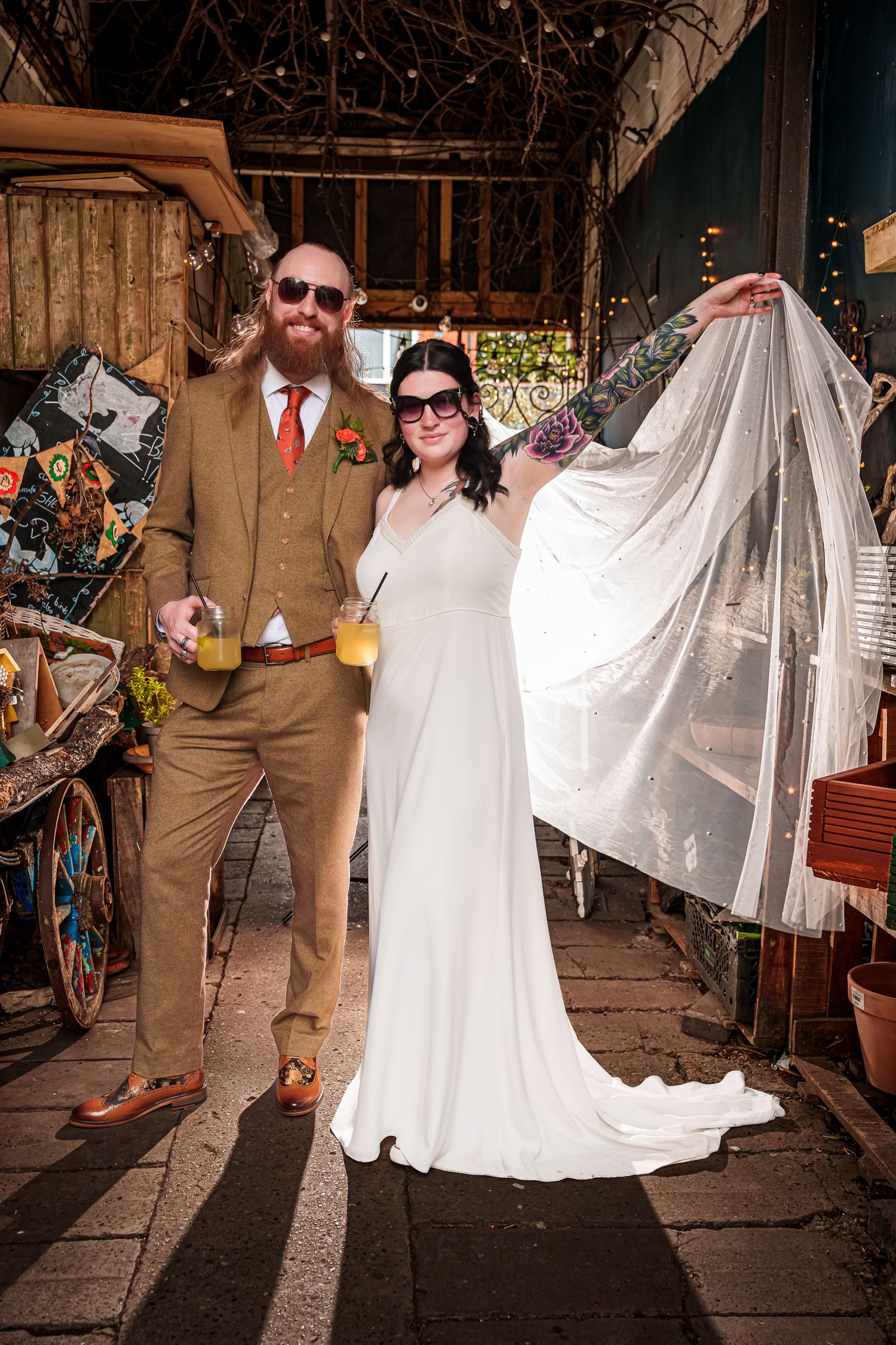 A bride in a white gown and groom in a tan suit hold drinks and pose together at a rustic outdoor wedding venue.