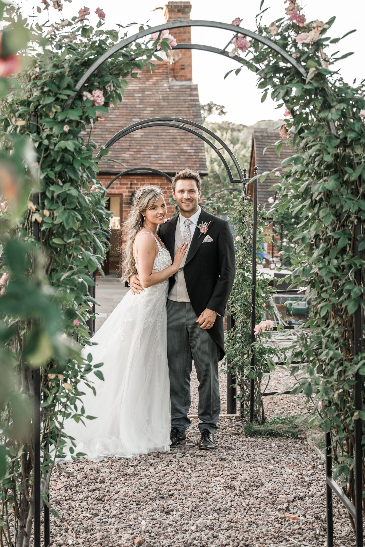 A romantic wedding archway made of green foliage and flowers frames a garden path with a brick building behind.
