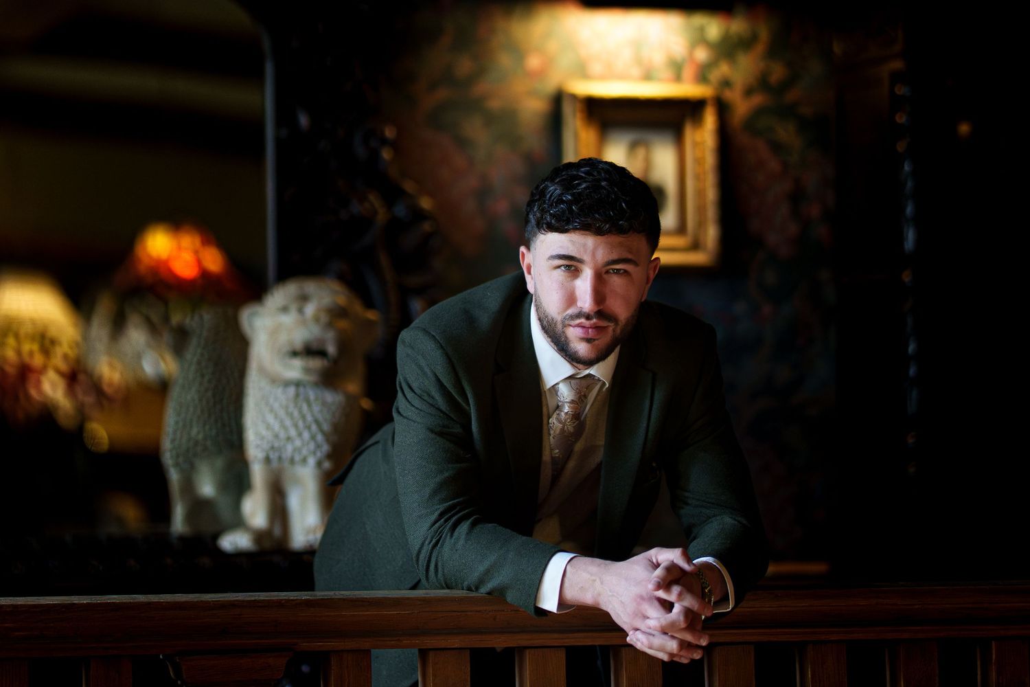 A young man in a dark green suit leans on a wooden railing in an elegant, dimly lit room with a stone lion statue.