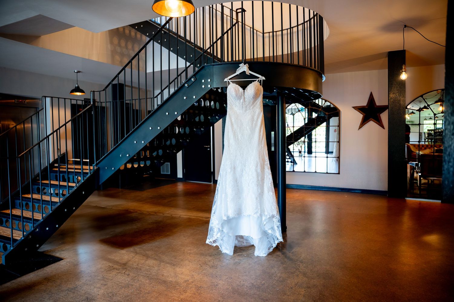 An elegant white wedding dress hangs in front of a spiral staircase in an industrial-chic space with rustic decor.