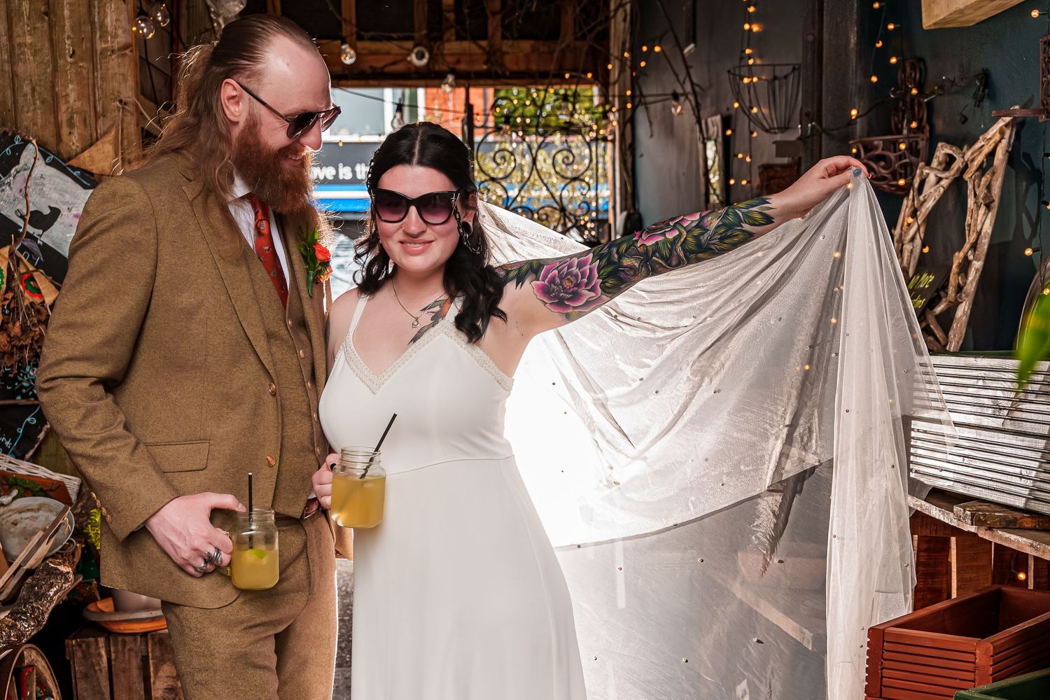 Bride in white gown with veil and groom in brown suit holding drinks at a rustic wedding venue.