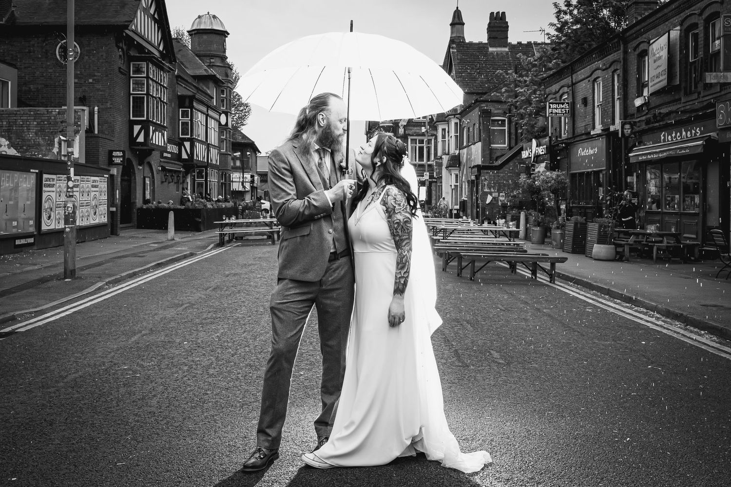 Black and white photo of a bride and groom sharing an umbrella on a street in a charming town.