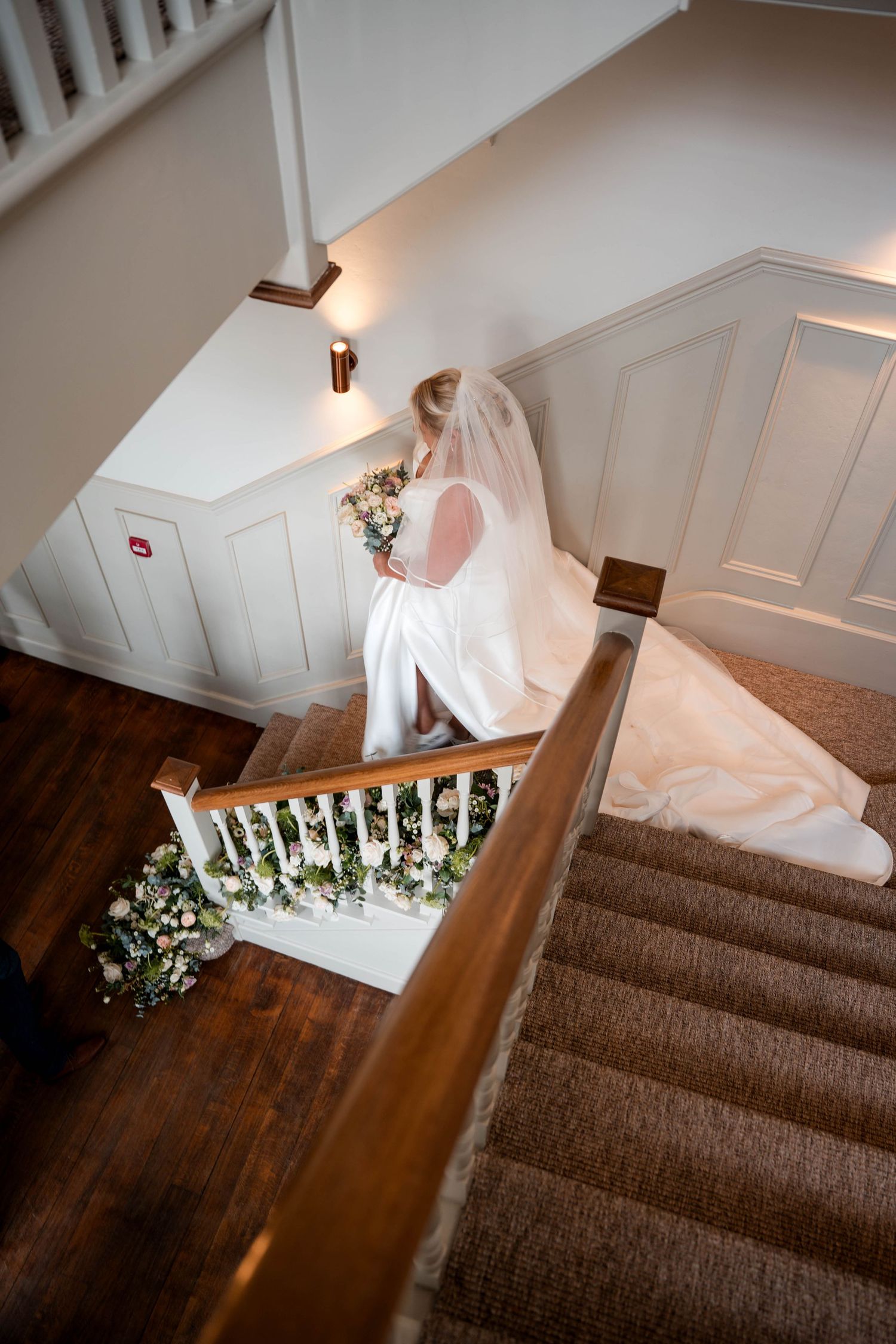 White wedding dress trailing down carpeted staircase decorated with floral arrangements.
Stockton house wedding venue